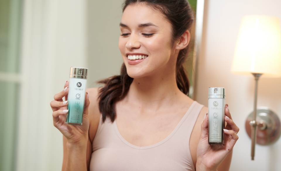 A photo of a woman holding up an Age IQ Day Cream bottle in her right hand and an Age IQ Night Cream bottle in her left hand.